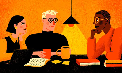 Three people sit at a table with notebooks, a laptop, and mugs, having a discussion under a hanging light.